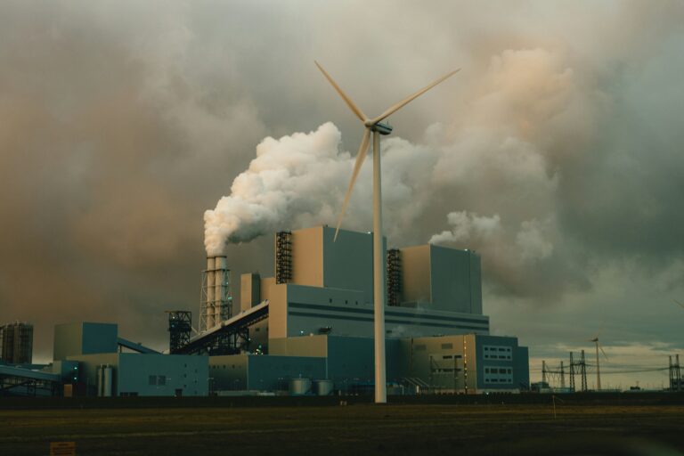 A wind turbine stands in front of a coal-burning power station that emits thick smoke into the air.